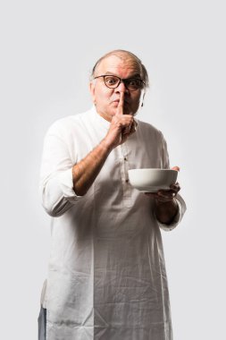 Yaşlı ya da yaşlı Hintli bir adam kaşıkla çatal kullanarak boş beyaz tabak ya da kâseden yemek yiyor. Beyaz arka planda farklı ifadelerle tek başına duruyor.