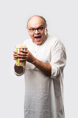 Beyaz arka planda patlamış mısır yiyen yaşlı, kel Hintli adam neşeli ifadeler takınıyor.