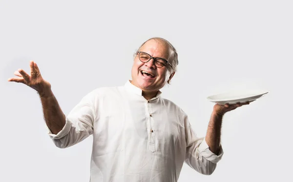 Senior or Old Indian asian man eating from empty white plate or bowl using spoon and fork - standing isolated over white background with different expressions