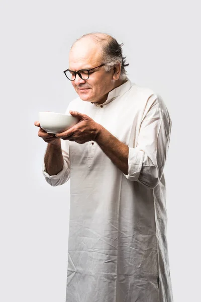 Senior or Old Indian asian man eating from empty white plate or bowl using spoon and fork - standing isolated over white background with different expressions