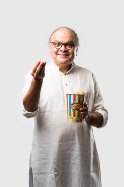 Beyaz arka planda patlamış mısır yiyen yaşlı, kel Hintli adam neşeli ifadeler takınıyor.