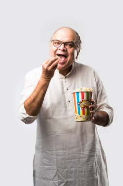 Beyaz arka planda patlamış mısır yiyen yaşlı, kel Hintli adam neşeli ifadeler takınıyor.