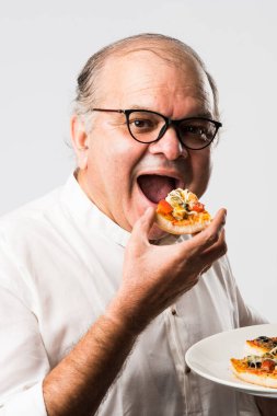 Beyaz arka planda tek başına dururken pizza yiyen yaşlı ya da yaşlı Hintli bir adam.