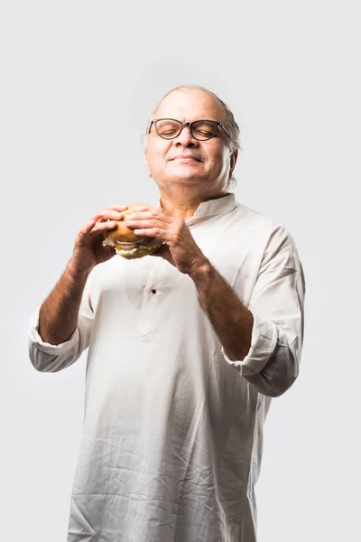 Indian Old Man eating burger or Hamburger with mango juice or cold drink