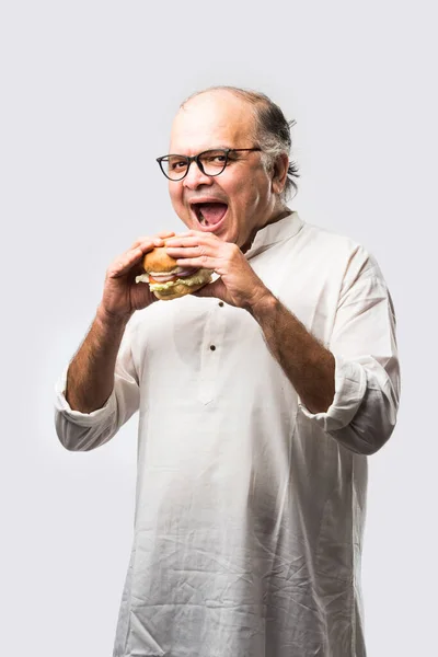Indian Old Man eating burger or Hamburger with mango juice or cold drink