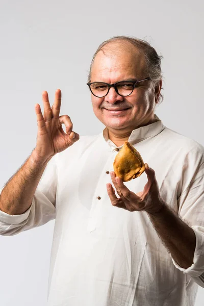 Indian old man or senior man eating samosa or veg pastry