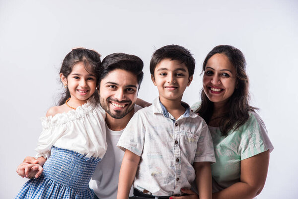Молодая индийская семья из четырех человек обнимается, смотрит в камеру и улыбается, стоя на белом фоне