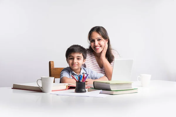 Cute Indian boy with mother doing homework at home using laptop and books - online schooling concept