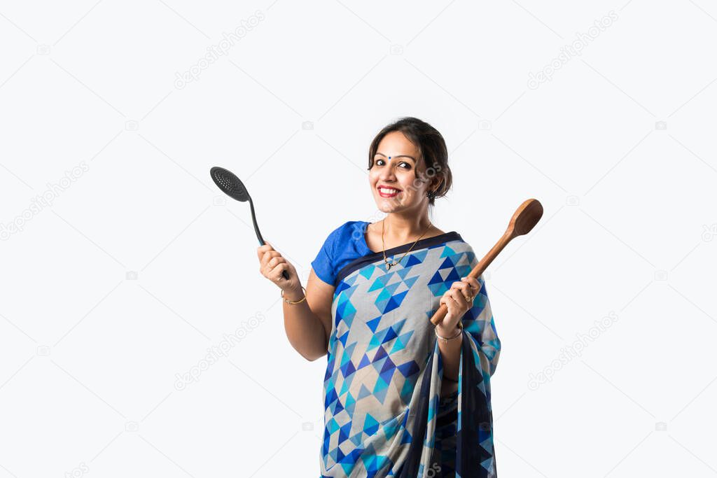 Portrait of an Indian asian woman in saree in kitchen, holding wooden ladle, belan, spatula or pan, isolated against white background