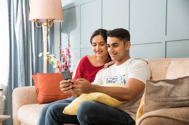 Hintli genç çift kanepe ya da kanepe ya da modern iç mekan ya da evlerinde otururken akıllı telefon kullanıyor.