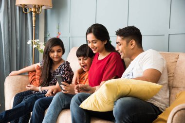 Hintli aile kanepede oturup akıllı telefon, dizüstü bilgisayar ya da tablet kullanıyor, film izliyor ya da internette sörf yapıyor.