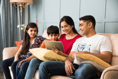 Hintli aile kanepede oturup akıllı telefon, dizüstü bilgisayar ya da tablet kullanıyor, film izliyor ya da internette sörf yapıyor.