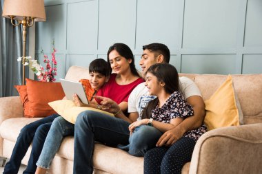 Hintli aile kanepede oturup akıllı telefon, dizüstü bilgisayar ya da tablet kullanıyor, film izliyor ya da internette sörf yapıyor.
