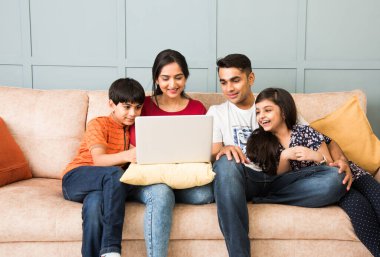 Hintli aile kanepede oturup akıllı telefon, dizüstü bilgisayar ya da tablet kullanıyor, film izliyor ya da internette sörf yapıyor.