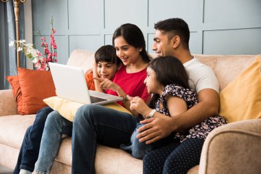 Hintli aile kanepede oturup akıllı telefon, dizüstü bilgisayar ya da tablet kullanıyor, film izliyor ya da internette sörf yapıyor.