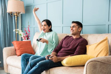 Hintli genç çift kanepede ya da kanepede otururken joystick ya da denetleyici kullanarak video oyunu oynuyorlar.