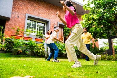 Ev bahçesinde göz kapağı oyunu oynayan Hintli aile, birlikte güzel vakit geçiriyorlar. Açık hava sporlarının tadını çıkarıyorlar. Ebeveynler ve çocuklar gülüyor, koşuyor ve oyuncu arka bahçe aktiviteleriyle kaynaşıyorlar.