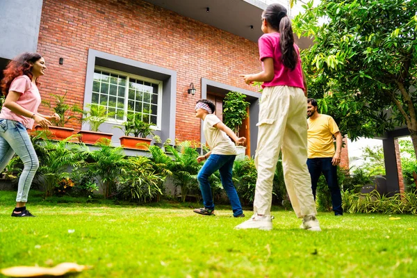 Ev bahçesinde göz kapağı oyunu oynayan Hintli aile, birlikte güzel vakit geçiriyorlar. Açık hava sporlarının tadını çıkarıyorlar. Ebeveynler ve çocuklar gülüyor, koşuyor ve oyuncu arka bahçe aktiviteleriyle kaynaşıyorlar.