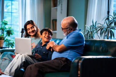 Büyükanne ve büyükbabası bilgisayar kullanan Hintli bir çocuk modern evdeki koltukta otururken, yaşlı büyükannesi ve büyükbabası çocuklara dijital öğrenme, aile bağı, teknoloji öğretiyor.
