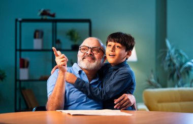 Hintli büyükbaba torununa evde sarılırken işaret ediyor, el hareketleriyle bir şeyler yapıyor, yaşlı bir adam torunuyla içeride oturuyor, iki insanın birbirine bağlandığı bir an.