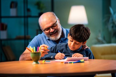 Hintli büyükbaba, çocuklara kalemle resim çizmede ya da boyamada yardım ediyor. İkisi de modern evdeki masada birlikte oturuyor. Yaşlı adam, torununa yaratıcı aktivitelerde rehberlik ediyor.