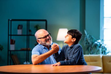 Hintli büyükbaba ve çocuk bilek güreşi yapıyorlar. İkisi de modern evdeki masada birlikte oturuyorlar. Yaşlı adam ve torun eğlenceli aktivitelerin keyfini çıkarıyorlar.