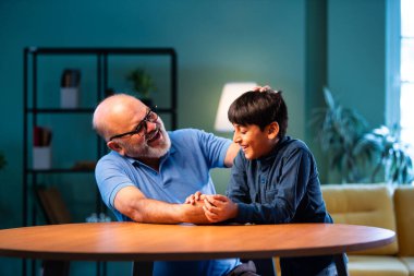 Hintli büyükbaba ve çocuk bilek güreşi yapıyorlar. İkisi de modern evdeki masada birlikte oturuyorlar. Yaşlı adam ve torun eğlenceli aktivitelerin keyfini çıkarıyorlar.