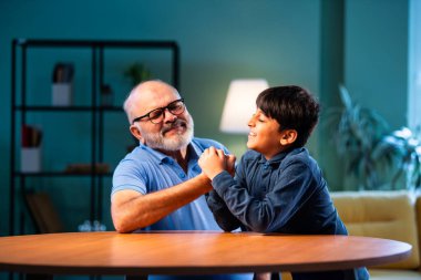 Hintli büyükbaba ve çocuk bilek güreşi yapıyorlar. İkisi de modern evdeki masada birlikte oturuyorlar. Yaşlı adam ve torun eğlenceli aktivitelerin keyfini çıkarıyorlar.