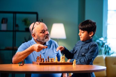 Hintli büyükbaba ve çocuk modern evde masada satranç oynuyorlar. Yaşlı büyükbabayla bağ kuruyorlar. Masa oyunlarından zevk alıyorlar.
