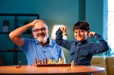 Hintli büyükbaba ve çocuk modern evde masada satranç oynuyorlar. Yaşlı büyükbabayla bağ kuruyorlar. Masa oyunlarından zevk alıyorlar.