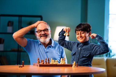 Hintli büyükbaba ve çocuk modern evde masada satranç oynuyorlar. Yaşlı büyükbabayla bağ kuruyorlar. Masa oyunlarından zevk alıyorlar.