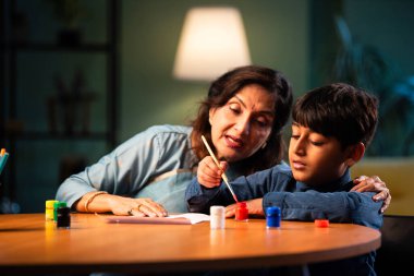 Çocuklu Hintli büyükanne, modern evdeki masada kırmızı renk ve fırçayla kalp boyuyor, yaşlı kadın çocuklara yaratıcı sanat aktiviteleri öğretiyor, sevgi sembolü üzerine bağ kuruyor, aile eğlencesi öğreniyor.