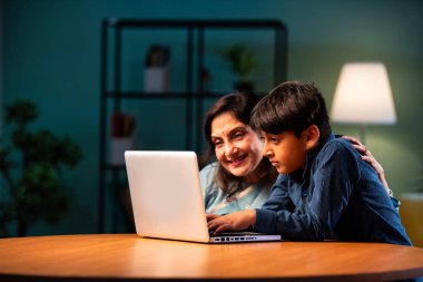 Çocuklu Hintli büyükanne modern evdeki masada dizüstü bilgisayar kullanıyor, yaşlı kadın çocuktan teknoloji öğreniyor, nesiller boyu aile bağı kuruyor, dijital eğitim, sevgi ve bakım.