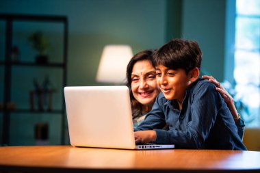 Çocuklu Hintli büyükanne modern evdeki masada dizüstü bilgisayar kullanıyor, yaşlı kadın çocuktan teknoloji öğreniyor, nesiller boyu aile bağı kuruyor, dijital eğitim, sevgi ve bakım.