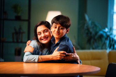 Evde büyükannesine sarılan Hintli çocuk. Büyükannesi masada otururken sevgi ve şefkat gösteriyor. Evde yaşlı bir kadınla aile bağı kuruyor.