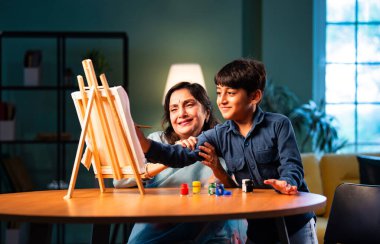 Çocuklu Hintli büyükanne, modern evdeki masada resim yapıyor, fırça ve renklerle çocuğa yol gösteriyor, aile bağı, yaratıcılık, sanat eğitimi, aşk ve eğlence.