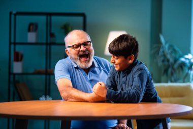 Hintli büyükbaba ve çocuk bilek güreşi yapıyorlar. İkisi de modern evdeki masada birlikte oturuyorlar. Yaşlı adam ve torun eğlenceli aktivitelerin keyfini çıkarıyorlar.