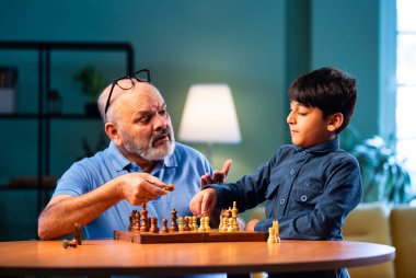 Hintli büyükbaba ve çocuk modern evde masada satranç oynuyorlar. Yaşlı büyükbabayla bağ kuruyorlar. Masa oyunlarından zevk alıyorlar.