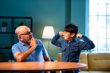 Hintli büyükbaba ve çocuk bilek güreşi yapıyorlar. İkisi de modern evdeki masada birlikte oturuyorlar. Yaşlı adam ve torun eğlenceli aktivitelerin keyfini çıkarıyorlar.