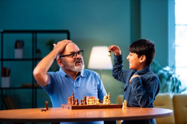 Hintli büyükbaba ve çocuk modern evde masada satranç oynuyorlar. Yaşlı büyükbabayla bağ kuruyorlar. Masa oyunlarından zevk alıyorlar.