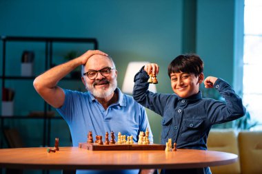 Hintli büyükbaba ve çocuk modern evde masada satranç oynuyorlar. Yaşlı büyükbabayla bağ kuruyorlar. Masa oyunlarından zevk alıyorlar.