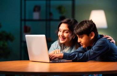 Çocuklu Hintli büyükanne modern evdeki masada dizüstü bilgisayar kullanıyor, yaşlı kadın çocuktan teknoloji öğreniyor, nesiller boyu aile bağı kuruyor, dijital eğitim, sevgi ve bakım.