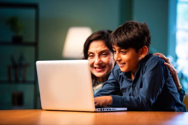 Çocuklu Hintli büyükanne modern evdeki masada dizüstü bilgisayar kullanıyor, yaşlı kadın çocuktan teknoloji öğreniyor, nesiller boyu aile bağı kuruyor, dijital eğitim, sevgi ve bakım.