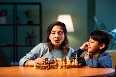 Hintli büyükanne ve çocuk modern evde masada satranç oynuyorlar, yaşlı kadın çocuklara stratejik oyun öğretiyor, aile bağı, zihinsel egzersiz, aşk, sabır, birliktelik