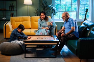 Hintli çocuk modern evde büyükanne ve büyükbabasıyla carrom oynuyor. Yaşlı büyükanne ve büyükbaba çocuklarla oyun oynuyor. Aile bağları, geleneksel Hint yaşam tarzı, eğlence, neşe.