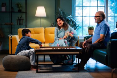 Hintli çocuk modern evde büyükanne ve büyükbabasıyla carrom oynuyor. Yaşlı büyükanne ve büyükbaba çocuklarla oyun oynuyor. Aile bağları, geleneksel Hint yaşam tarzı, eğlence, neşe.