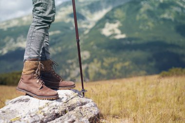 Kahverengi deri çizmeleri ve yürüyüş sopasıyla arka planda dağları olan bir kayanın üzerinde duran kadının yakın plan detayları.
