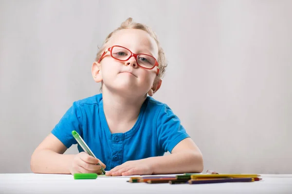 The boy wears glasses, sits at the table and draws.