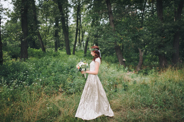 Elegant bride with wreath on head in light cream wedding dress standing in summer forest.