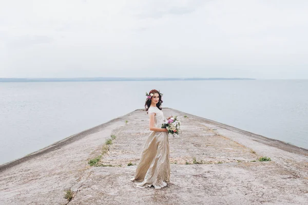 Bride with wreath and bouquet in hands standing against river background.